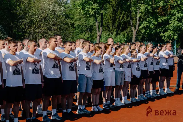 Group of young people in white T-shirts standing in rows on an outdoor track, arms folded, facing left.