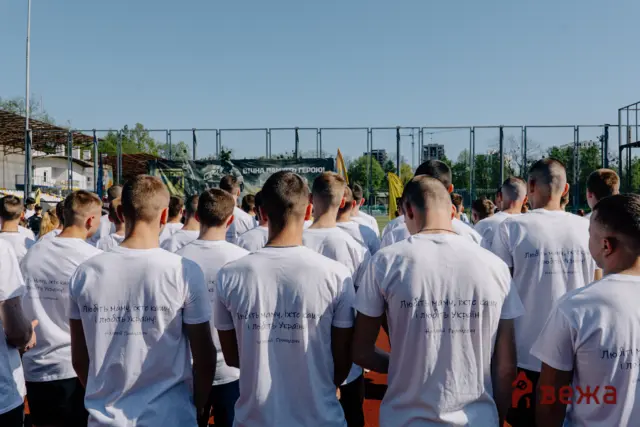 Crowd of people in white T-shirts standing shoulder to shoulder on a sports field, viewed from behind, with a chain-link fence and a banner in the background.