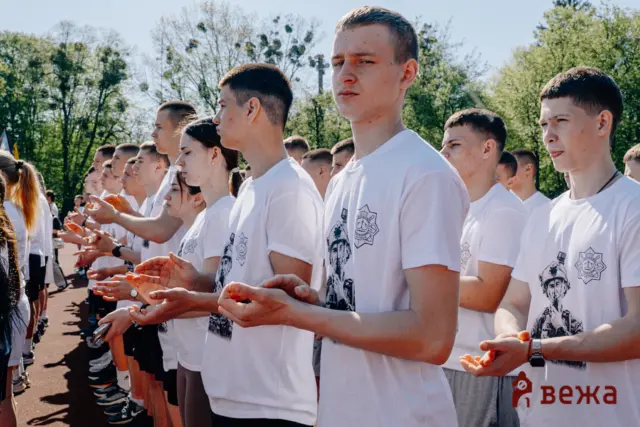 Group of young people in white T-shirts standing in a row outdoors, clapping during a ceremony or event.