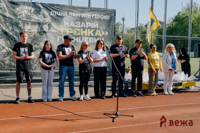 Group of people standing on a track in front of a large banner with a soldier image, clapping during a ceremony.