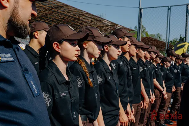 Line of uniformed cadets standing at attention outdoors, wearing brown caps and dark shirts with badges, facing left.