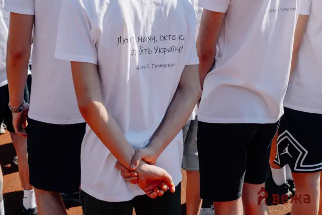 Back view of a group wearing white T-shirts, standing with hands clasped in front; one shirt has Cyrillic writing.