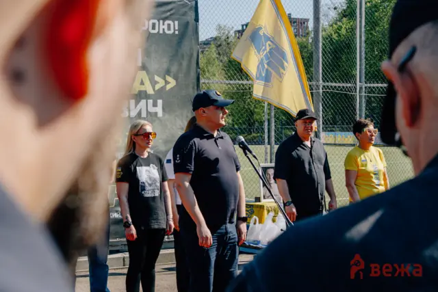 Group of people standing outdoors at a fenced event, one person speaks into a microphone while a yellow flag waves in the background.
