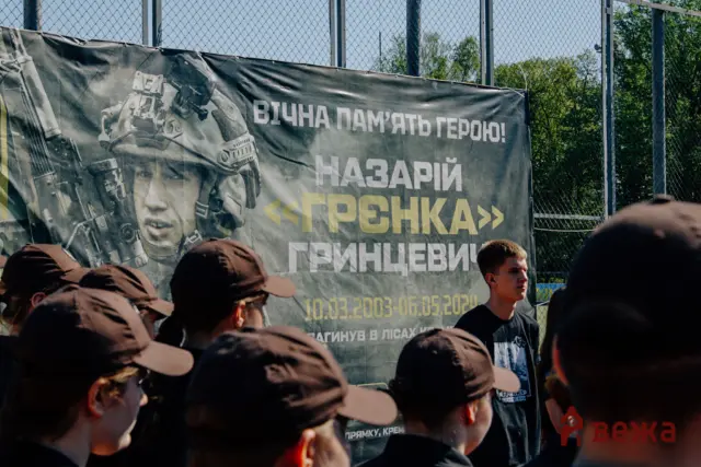 Banner featuring a soldier in combat gear with bold Cyrillic text, displayed behind a crowd at an outdoor fenced area.