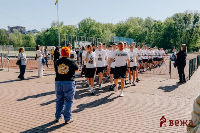 Group of runners in white shirts walking along a track as spectators photograph them on a sunny day, with trees and fencing in the background.