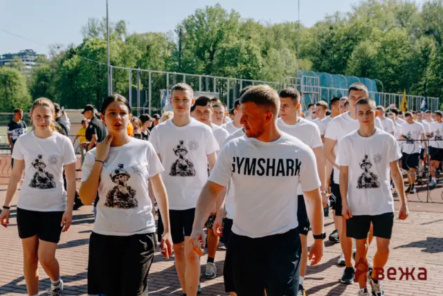 Group of young people in white T-shirts marching in formation on a sunny outdoor sports field.