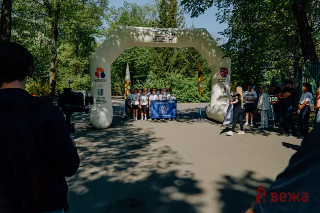 Group of people in white shirts under a white inflatable arch at an outdoor event among trees, with banners and flags nearby.