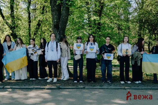 Group of young people standing in a park holding signs with blue hearts and a Ukrainian flag visible in the foreground.