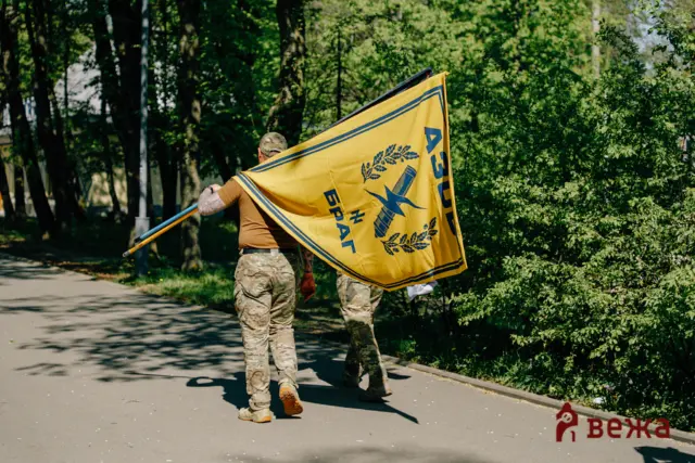 Soldier in camouflage holding a large yellow flag with a blue emblem outdoors among trees.