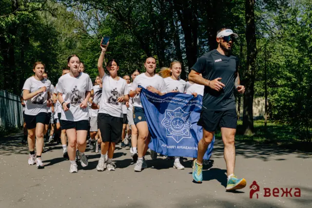 Group of runners in white shirts sprinting along a sunlit park path, a man in front with a dark shirt and cap holding a blue banner with an emblem.