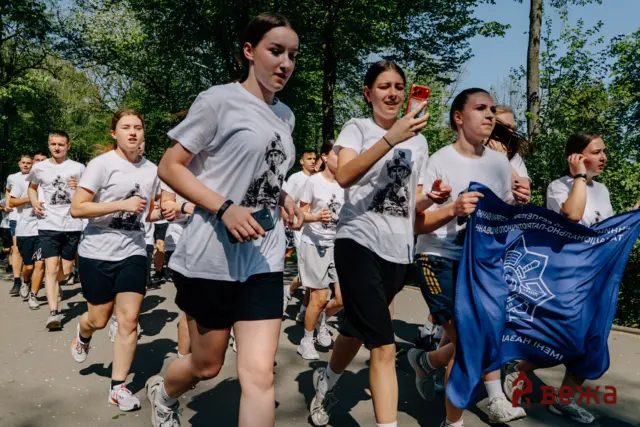 Group of young runners wearing white T-shirts sprint along a tree-lined park path during a race.