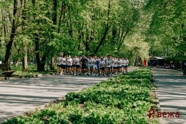 A large group of runners in white shirts jog along a tree-lined park path on a sunny day, with green shrubs in the foreground.
