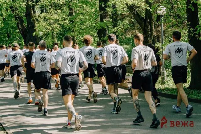Group of runners wearing matching gray T-shirts with a shield logo, jogging along a tree-lined park path.