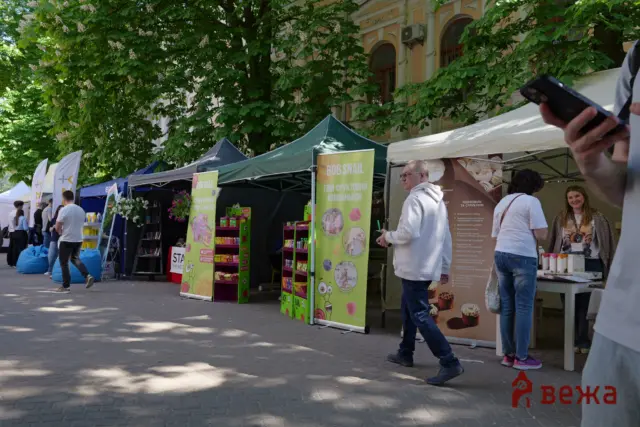 Outdoor market with colorful vendor tents lining a sidewalk and people browsing under leafy trees