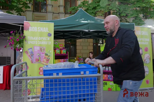 Man pushing a blue recycling bin in front of a bright market stall with green banners reading BOB SNAIL