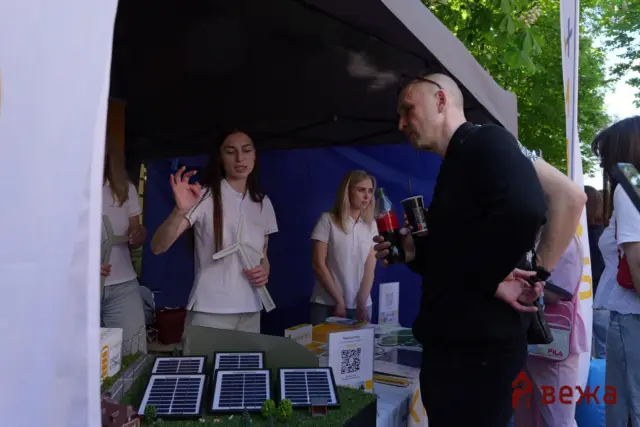 Staff and visitors at an outdoor booth showcasing solar panel models on a display table under a canopy.
