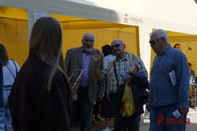 Group of adults conversing at an outdoor event under a yellow tent; one man in a blazer waves his hand at the camera.