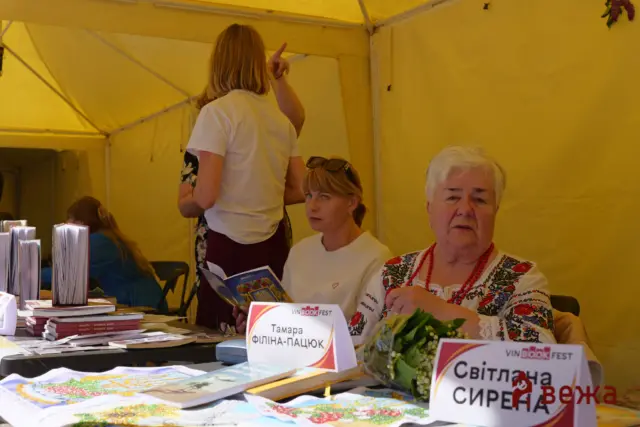 Book stall at VinBook Fest under a yellow tent; a seated woman reads a brochure while another woman stands with her back to the camera.