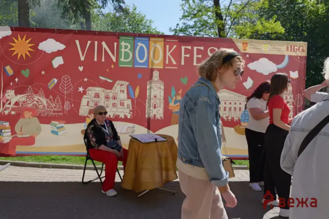 Outdoor book festival scene with a large red banner, people standing, and a seated woman signing books at a table.