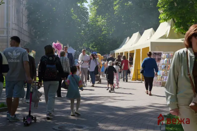 Crowd walking along a street market with white tents and booksellers stalls on the right under trees in sunlight.
