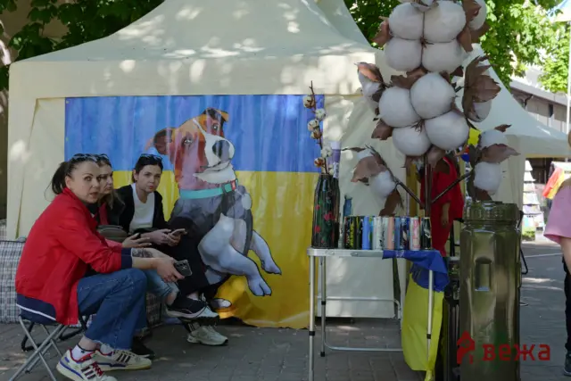 Four women sit at an outdoor market booth under a white canopy, with a blue-yellow dog banner behind them and a cotton‑candy display to the right.
