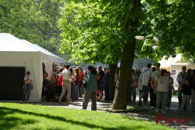 Crowd of people at an outdoor park fair with white vendor tents under leafy trees on a sunny day.