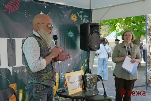 Older man with a white beard speaks into a handheld microphone at an outdoor booth, wearing a patterned vest; a woman with papers stands nearby.