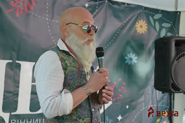 Bearded man in a patterned vest speaking into a handheld microphone on a stage with floral banner backdrop.