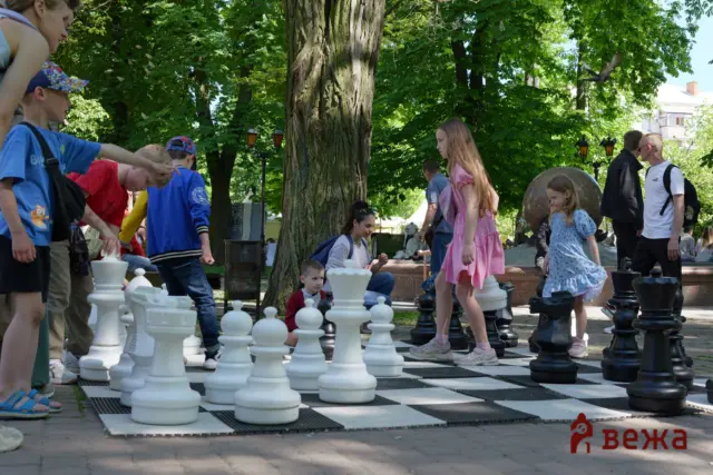 Children and adults playing a giant outdoor chess game on a checkered board in a sunny park with trees nearby.