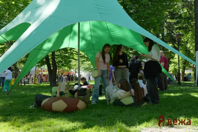 Group of people relaxing under a large teal canopy in a sunny park.