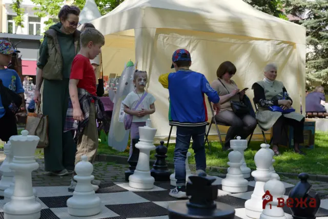 Group of people, including children, gathered around a life-size outdoor chessboard with large white and black pieces near a beige tent park-side.