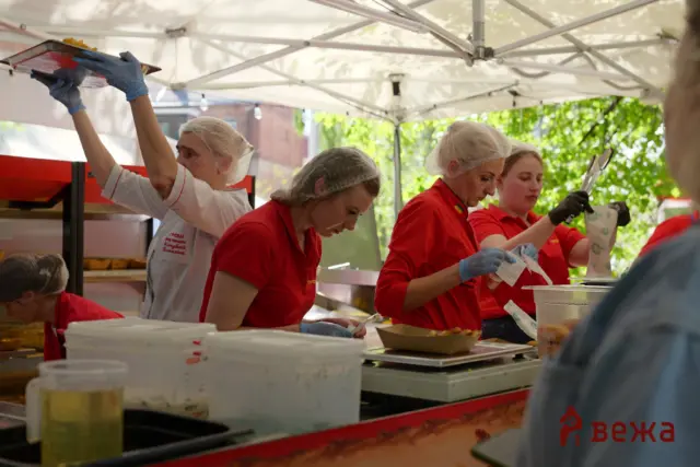 Staff in hairnets and gloves prepare and serve food at a market stall under a canopy, with red uniforms and toppings ready on the counter