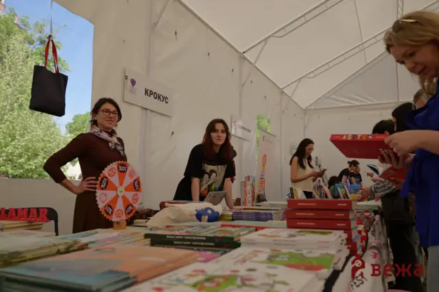 People at a book fair booth under a white tent, table piled with colorful books and a spinning prize wheel in the foreground.