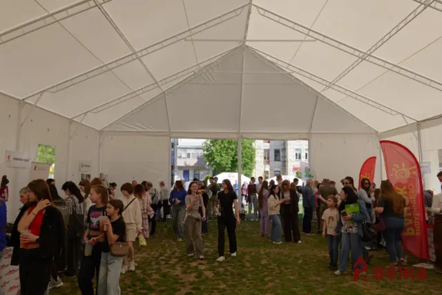 Crowd of people inside a large white event tent with banners and red flags, city buildings visible outside the tent entrance.