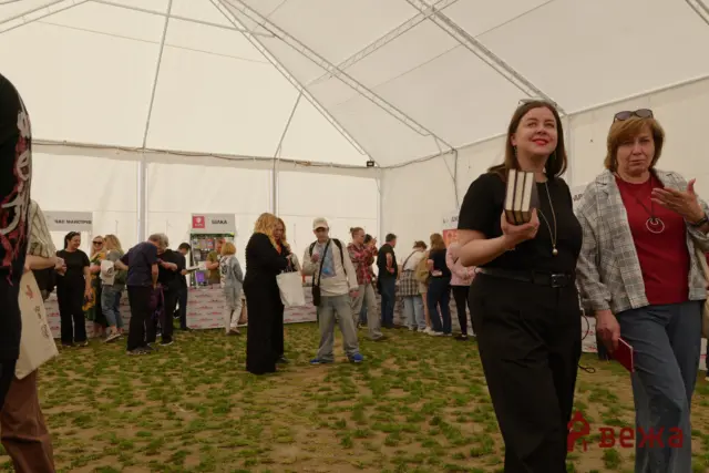 Two women stand at the edge of a busy indoor festival tent, one holding several books in her hand.