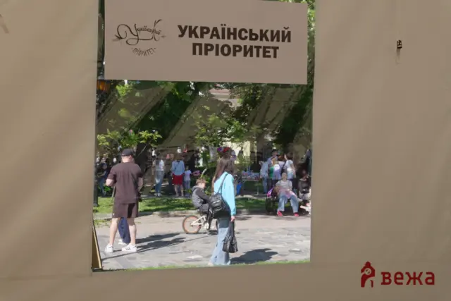 Group of people in a park seen through a rectangular opening in a wall; a beige banner overhead reads 'Український пріоритет' (Ukrainian Priority).
