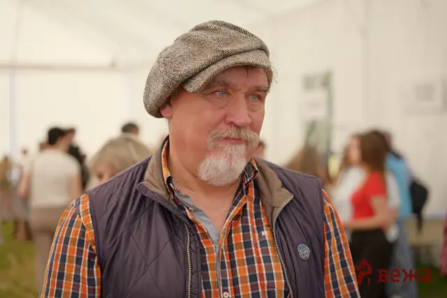 Close-up of an older man with a beige patterned beret, plaid shirt, and vest at an outdoor event with people in the background