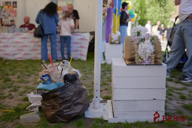 Outdoor festival scene with a booth in the background and a trash pile in the foreground; a stack of folded book-like panels with white and purple flowers sits on a white pedestal.