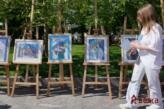 Outdoor art exhibit with framed paintings on wooden easels in a sunny park; a person in white holds a brochure on the right edge.