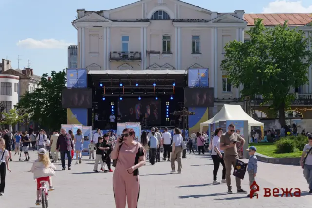 Outdoor city festival with a large stage and banners in front of a historic building under a blue sky, crowd watching a performance