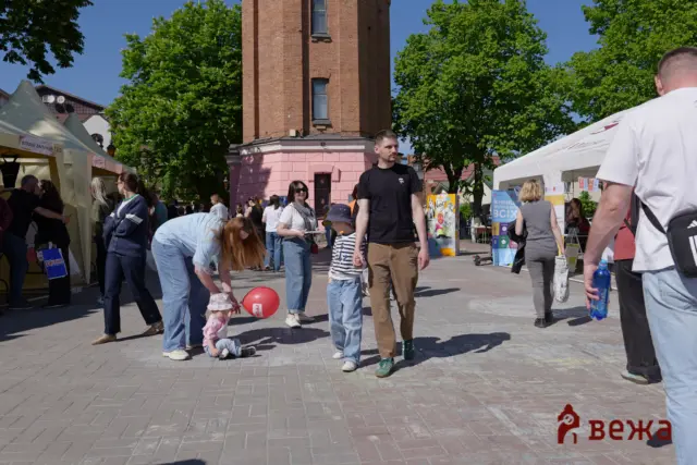 Crowd at an outdoor market with white tents, a brick tower in the background, and a child with a red balloon on the pavement.