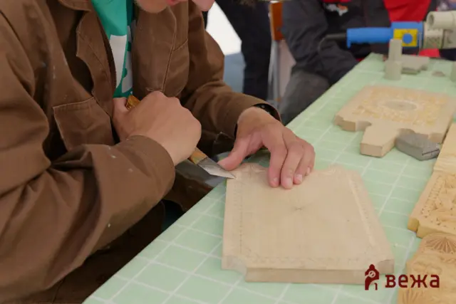 Person in a brown jacket carving a wooden block with a knife at a crafts table, with patterned wooden pieces nearby.