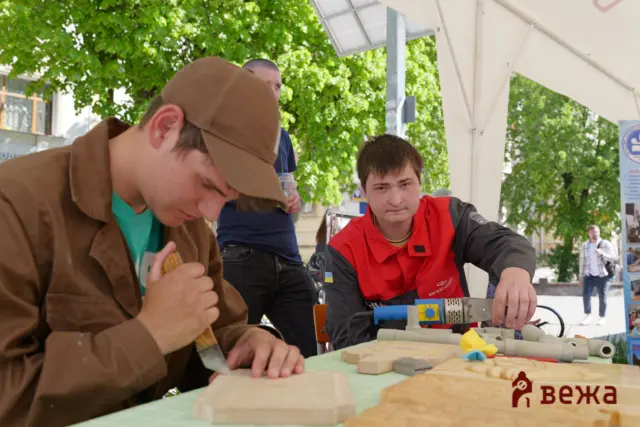 Two young men sit under a white tent outdoors, working with wood pieces and small tools on a table with a green mat and a workshop setup behind them.