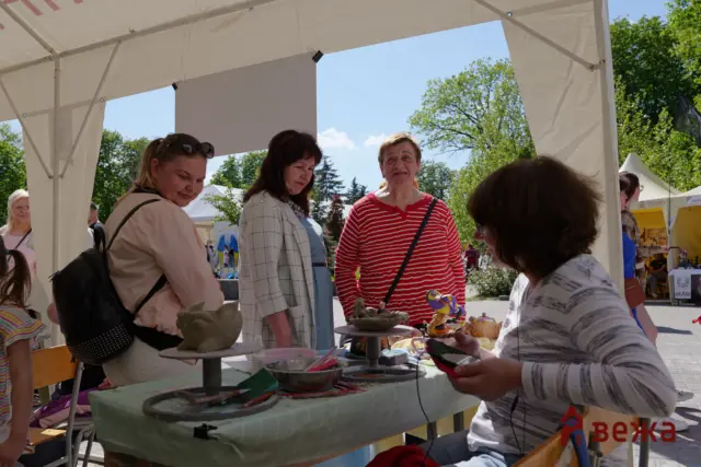 Group of people browsing handmade clay figurines at an outdoor market stall under a white canopy on a sunny day.