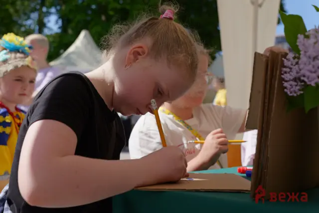 Girl in a black shirt leaning over a table and drawing with a pencil at an outdoor craft booth. The scene includes other children and festival tents in the background.