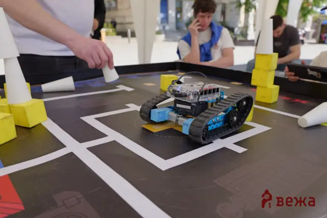Small wheeled robot on a dark competition mat with white lane markings, spectators in the background.
