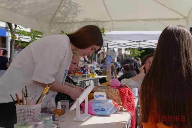 People at an outdoor craft fair, a woman leaning over a table of art supplies under a white canopy, helping a child with a project.