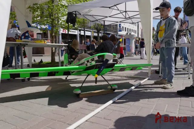 Green and black RC airplane on a display stand at an outdoor event, with tents and people in the background.