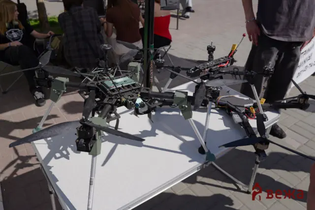 Quadcopter drones with exposed electronics placed on a white table at an outdoor event, people in the background observing.
