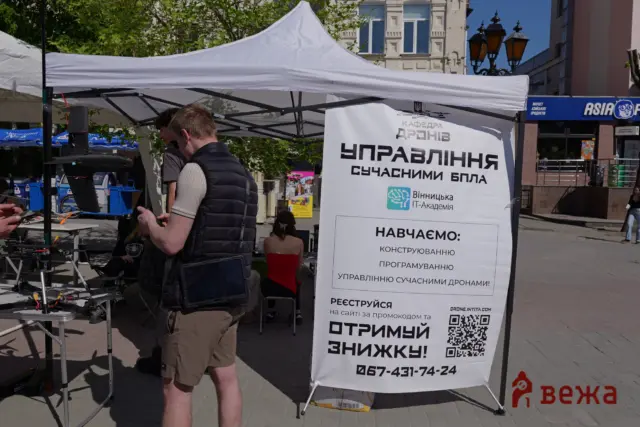 Outdoor drone booth under a white canopy with people handling drone gear and a large Ukrainian sign promoting IT Academy and modern drone management.